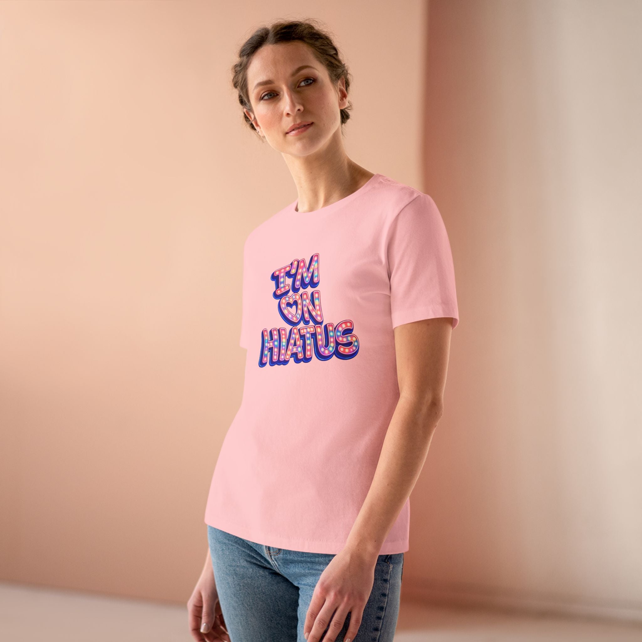 A woman wearing a light pink crewneck t-shirt with a colorful graphic that reads 'I'M ON HATUS' in a retro bubble font, paired with blue jeans, set against a warm beige studio background.