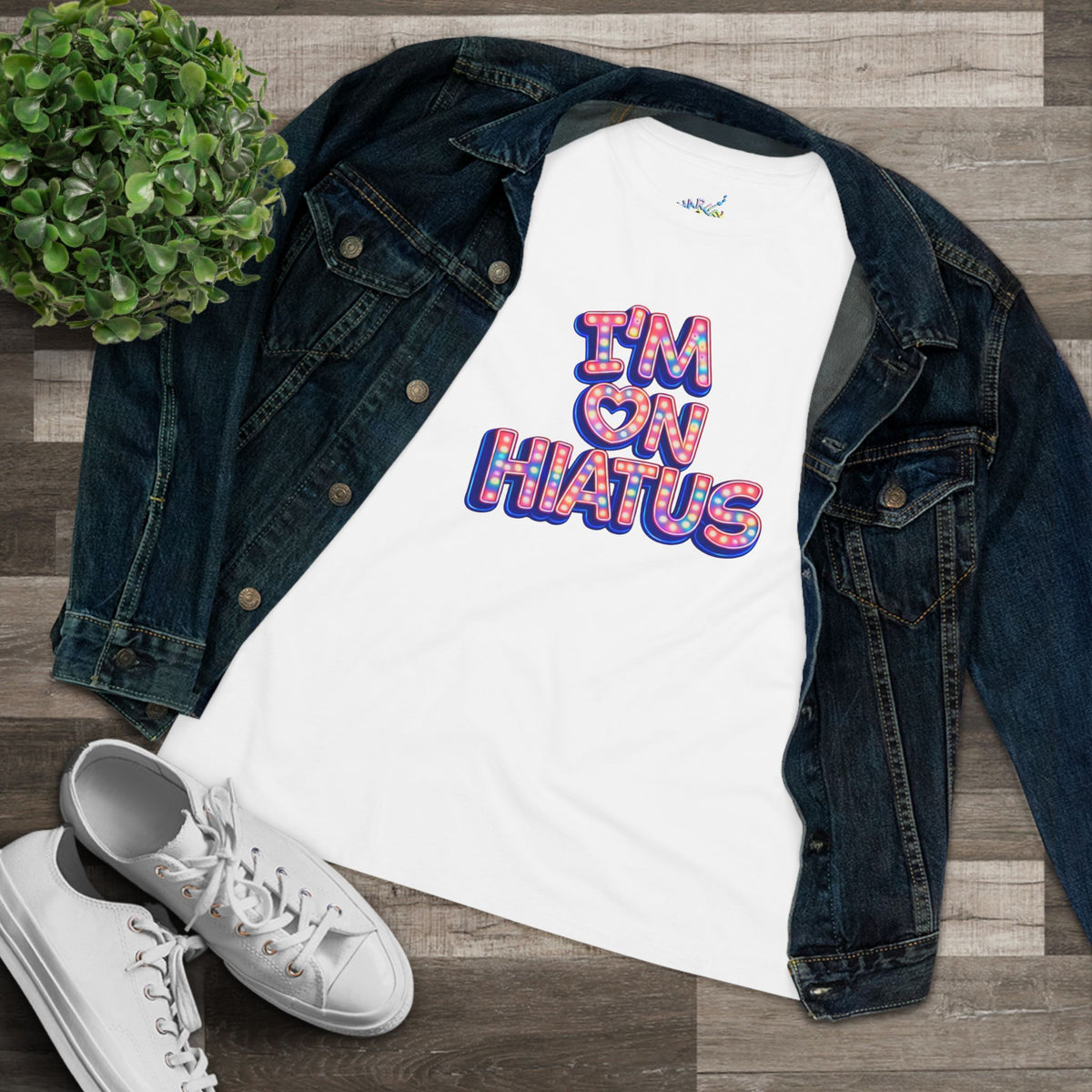 A white short-sleeve graphic t-shirt with a colorful retro-style marquee design reading 'I'M ON HIATUS', styled in a flat lay with a dark denim jacket, white sneakers, and a small green plant.