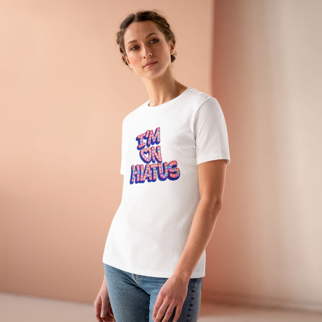 A woman modeling a white short-sleeve crewneck t-shirt with the colorful retro marquee "I'M ON HIATUS" graphic on the front, paired with blue jeans, set against a warm beige studio background.