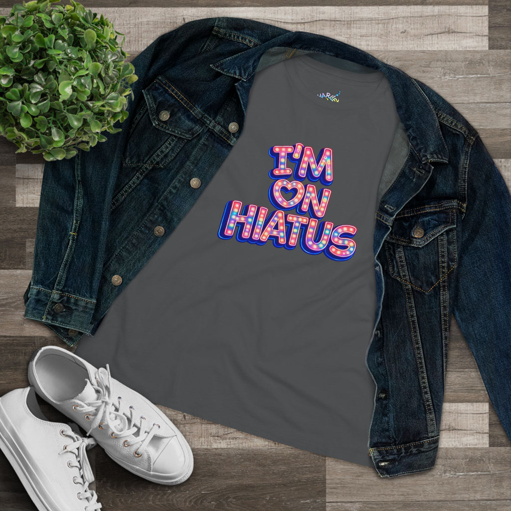 A dark gray graphic t-shirt with the text 'I'M ON HIATUS' in a colorful retro bubble font, styled in a flat lay with an open blue denim jacket, white sneakers, and a small green plant on a wood plank background.