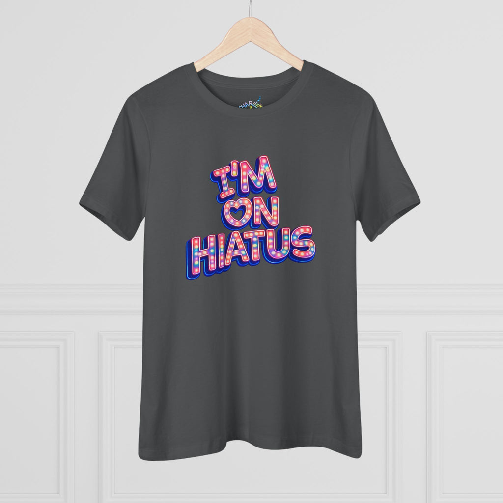 A dark gray crewneck t-shirt hanging on a wooden hanger against a white paneled wall. The shirt features a graphic text print reading 'I'M ON HIATUS' in a 3D bubble font filled with a red, white, pink, and blue pattern.