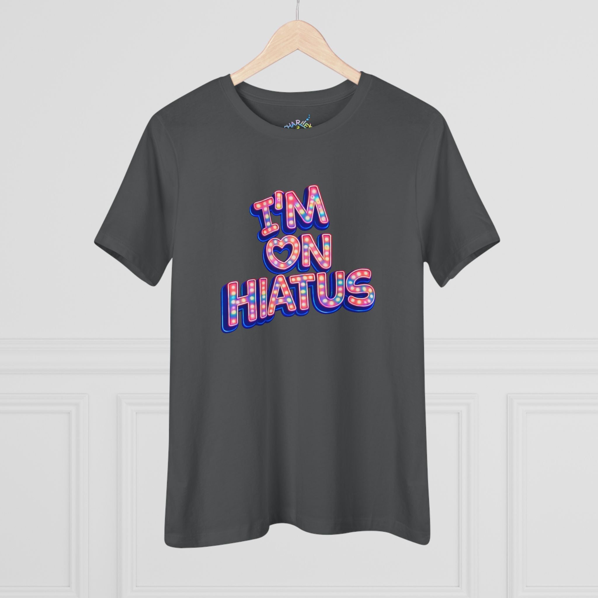 A dark gray crewneck t-shirt hanging on a wooden hanger against a white paneled wall. The shirt features a graphic text print reading 'I'M ON HIATUS' in a 3D bubble font filled with a red, white, pink, and blue pattern.