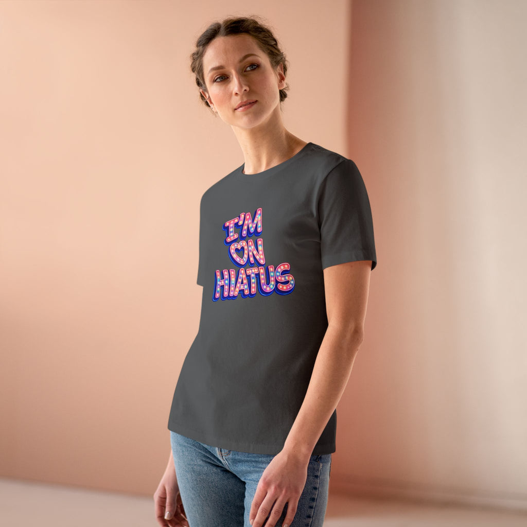 A woman wearing a dark gray t-shirt with a colorful graphic that reads 'I'M ON HIATUS' in pink, purple, and blue bubble letters, paired with blue jeans, set against a soft peach-colored studio background.