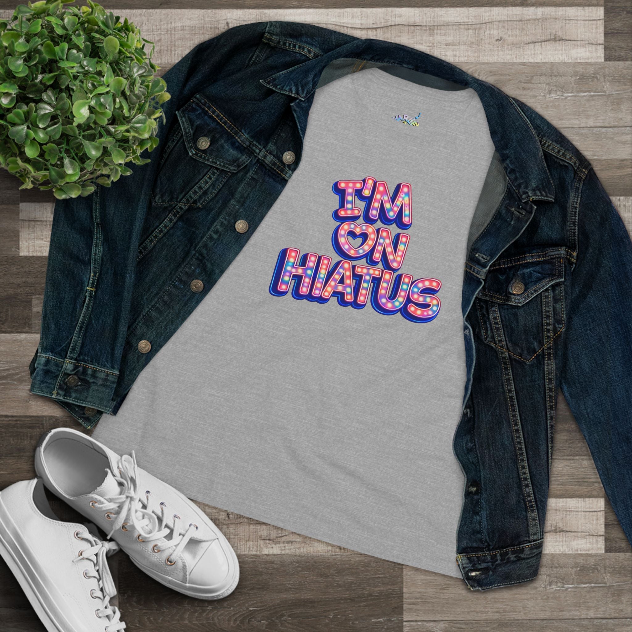 A light gray graphic t-shirt with a retro-style 'I'M ON HIATUS' design in red, white, and blue, styled in a flat lay with a dark blue denim jacket, white sneakers, and a green potted plant on a wood plank background.