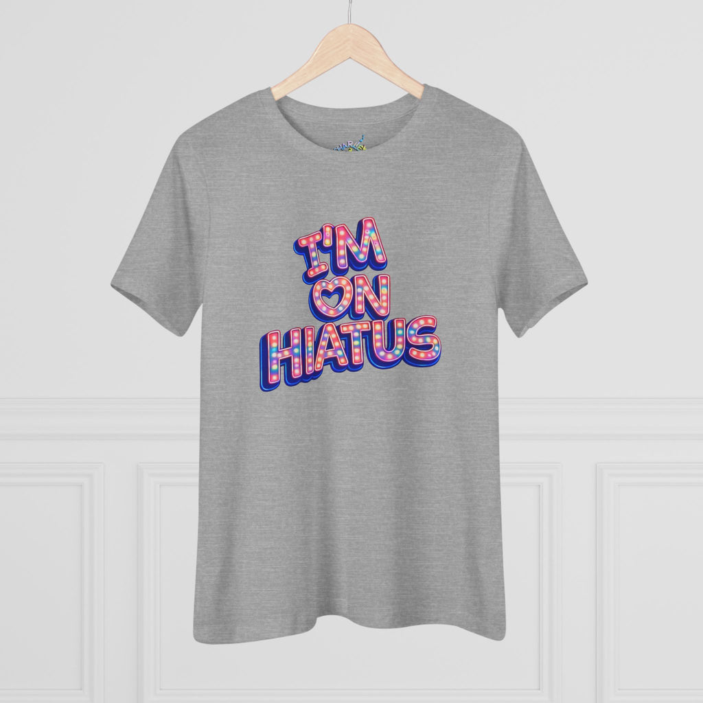 A heather gray short-sleeve t-shirt hanging on a wooden hanger against a white paneled wall. The shirt features a graphic text print reading 'I'M ON HIATUS' in a 3D block font filled with a red, white, pink, and blue pattern.