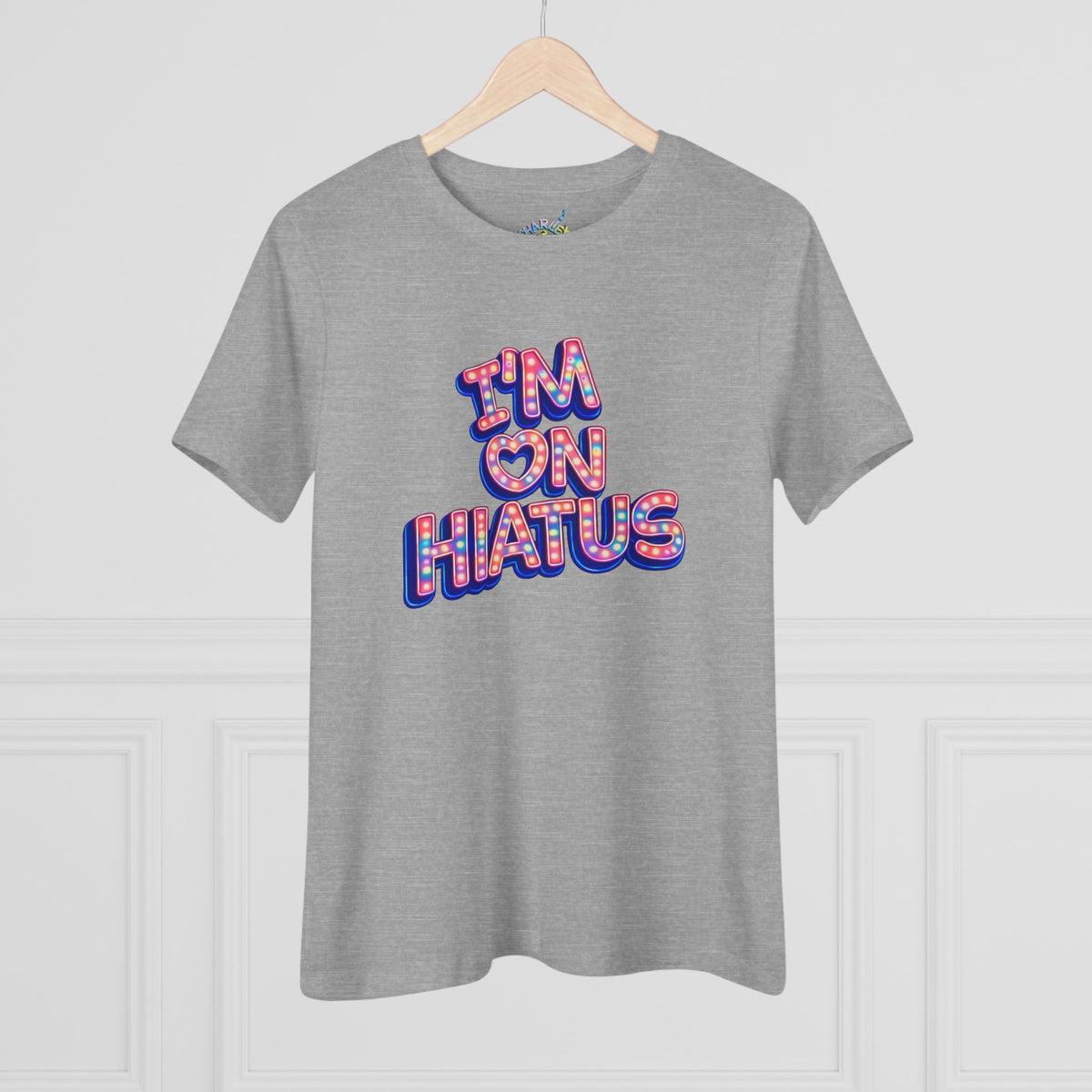 A heather gray short-sleeve t-shirt hanging on a wooden hanger against a white paneled wall. The shirt features a graphic text print reading 'I'M ON HIATUS' in a 3D block font filled with a red, white, pink, and blue pattern.