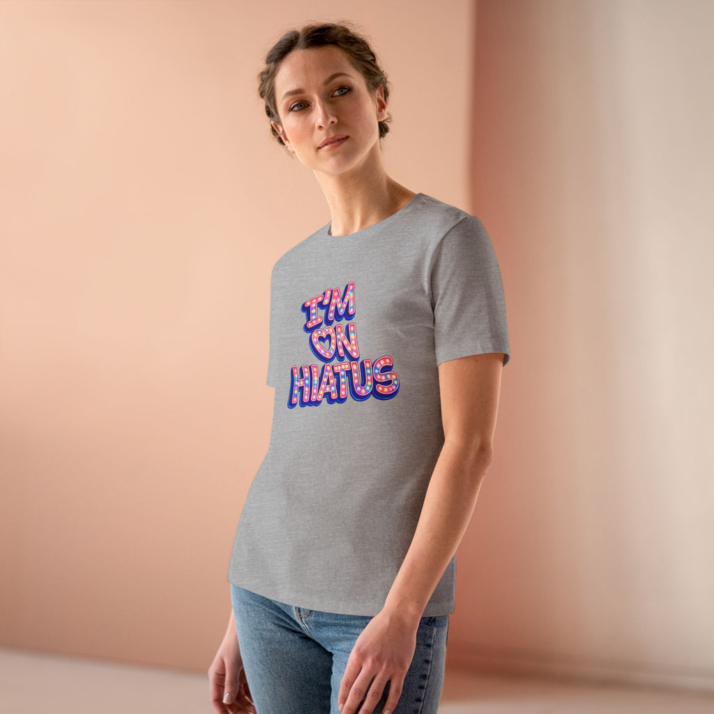 A woman wearing a heather gray t-shirt with a colorful retro-style graphic that reads 'I'M ON HIATUS' in pink, purple, and blue bubble letters, paired with blue jeans, set against a warm peach-colored studio background.