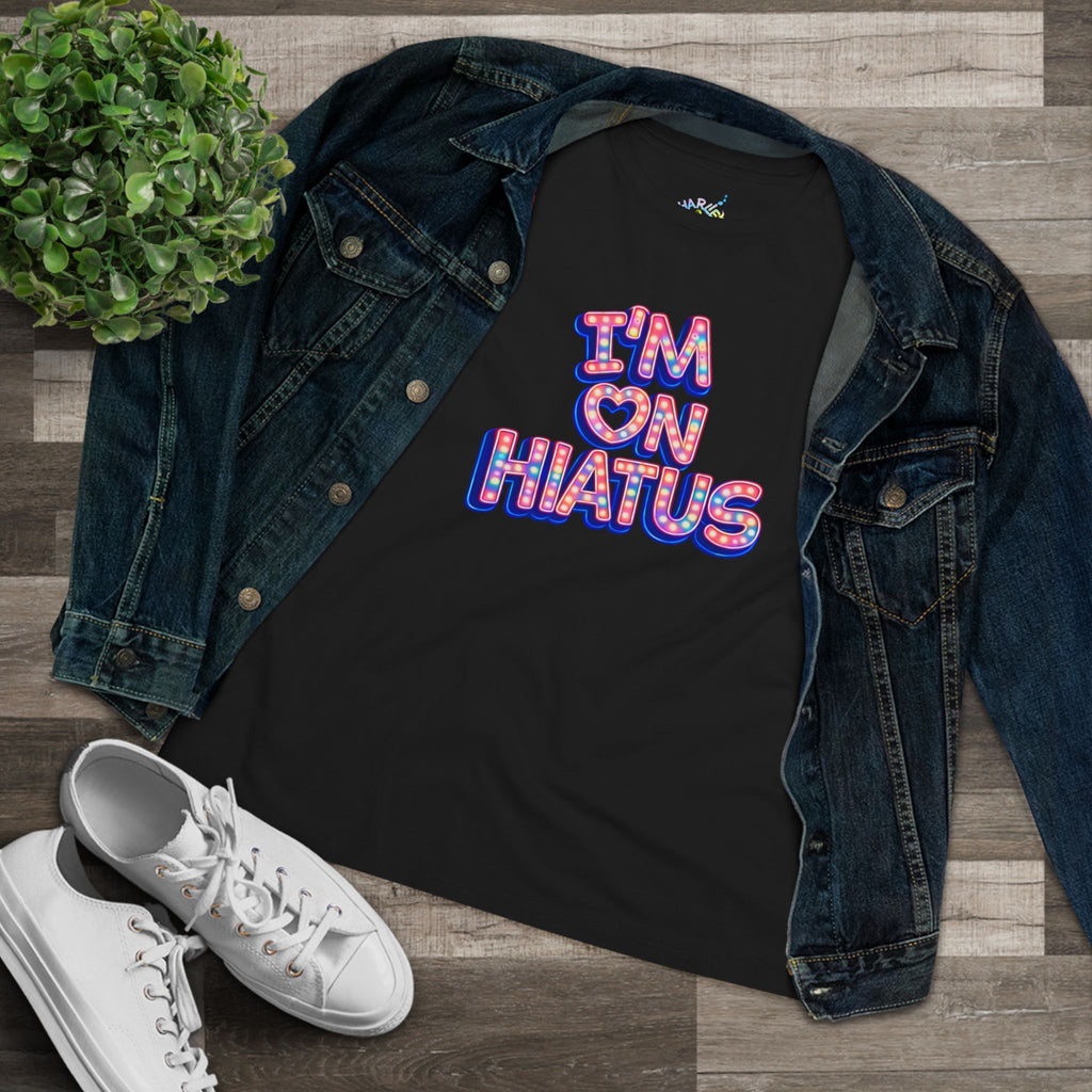A black graphic t-shirt with the text 'I'M ON HIATUS' in a colorful retro bubble font, styled in a flat lay with an open blue denim jacket, white sneakers, and a small green plant on a wood plank background.