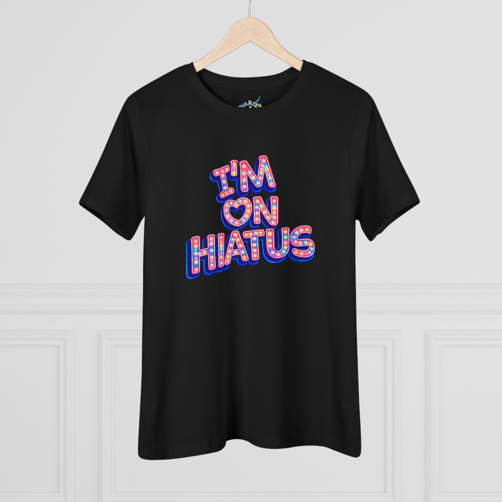 A black short-sleeve t-shirt hanging on a light wood hanger against a white paneled wall. The shirt features a colorful graphic on the chest reading 'I'M ON HIATUS' in a bold, 3D-effect font filled with a red, white, pink, and blue pattern.