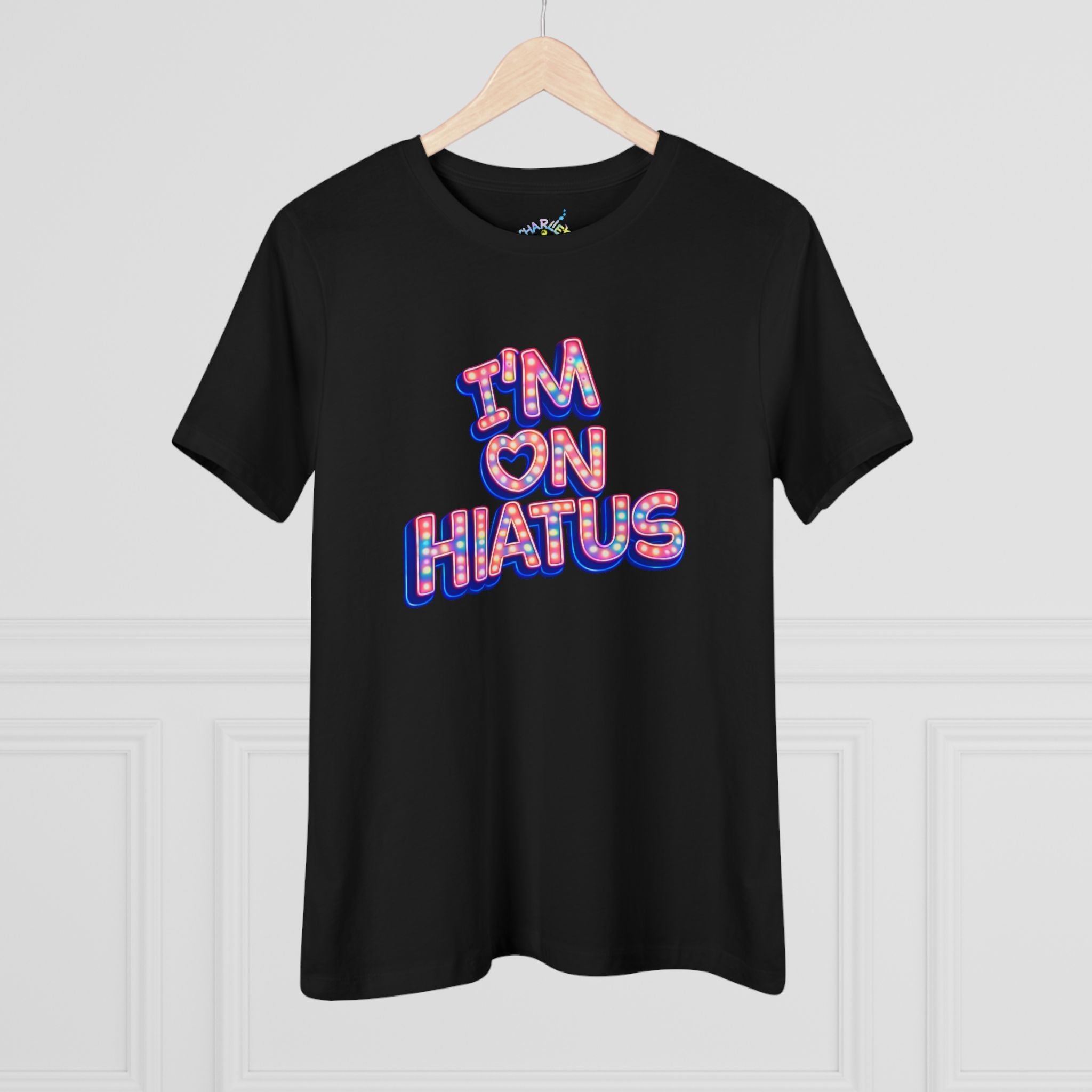 A black short-sleeve t-shirt hanging on a light wood hanger against a white paneled wall. The shirt features a colorful graphic on the chest reading 'I'M ON HIATUS' in a bold, 3D-effect font filled with a red, white, pink, and blue pattern.