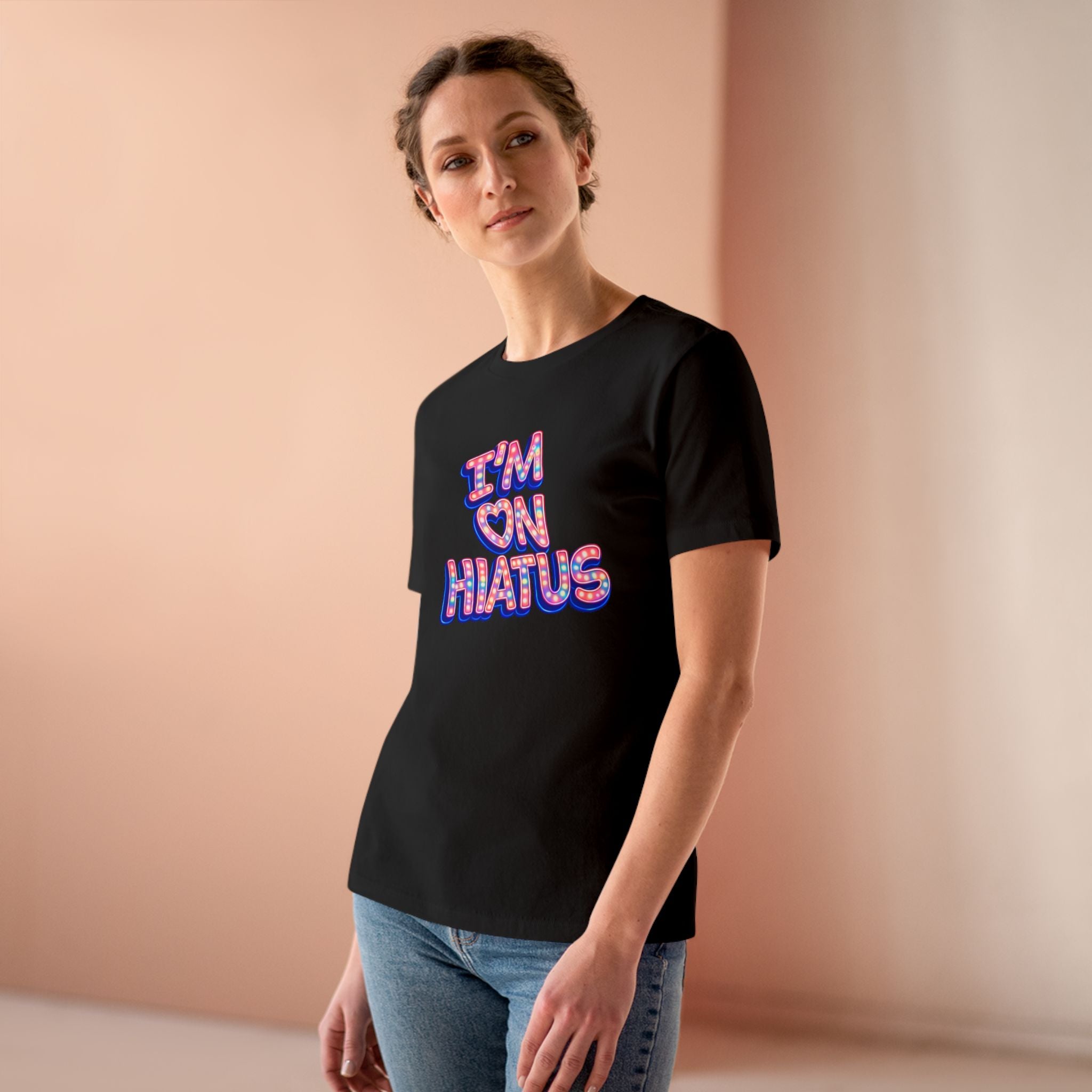 A woman wearing a black t-shirt with a colorful 'I'M ON HIATUS' graphic, paired with blue jeans, standing against a soft peach-colored background.