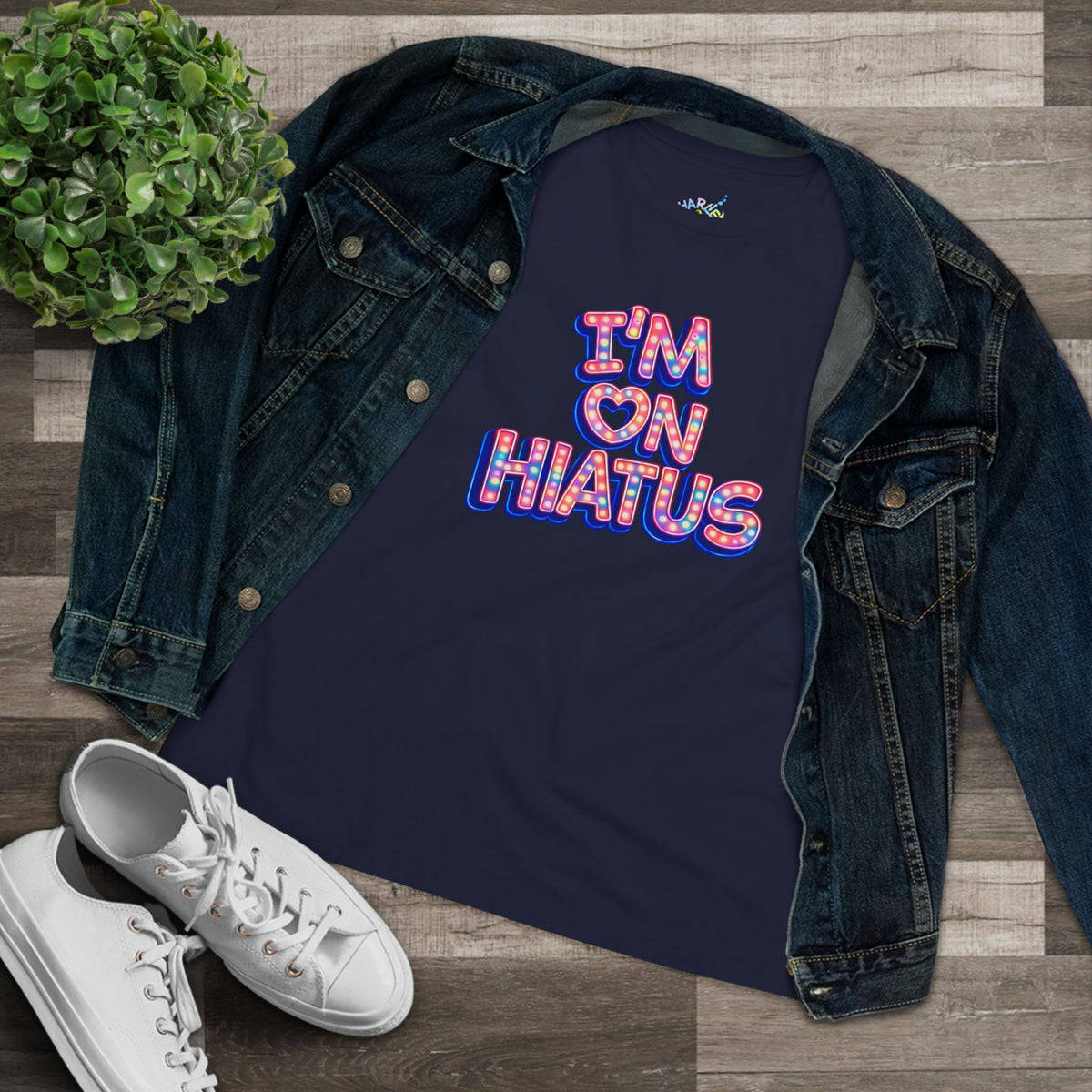 A navy blue graphic tee with the text 'I'M ON HIATUS' in a colorful retro bubble font, styled in a flat lay with an open dark denim jacket, white sneakers, and a small green plant on a wood plank background.