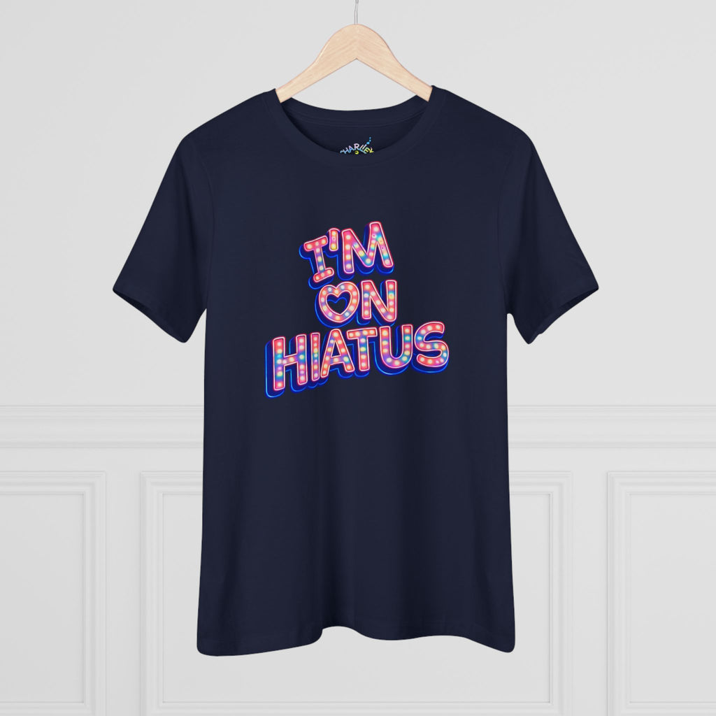 A navy blue short-sleeve t-shirt hanging on a light wood hanger against a white paneled wall. The shirt features a graphic text print reading 'I'M ON HIATUS' in a 3D bubble font filled with a red, white, pink,  and blue pattern and outlined in blue.
