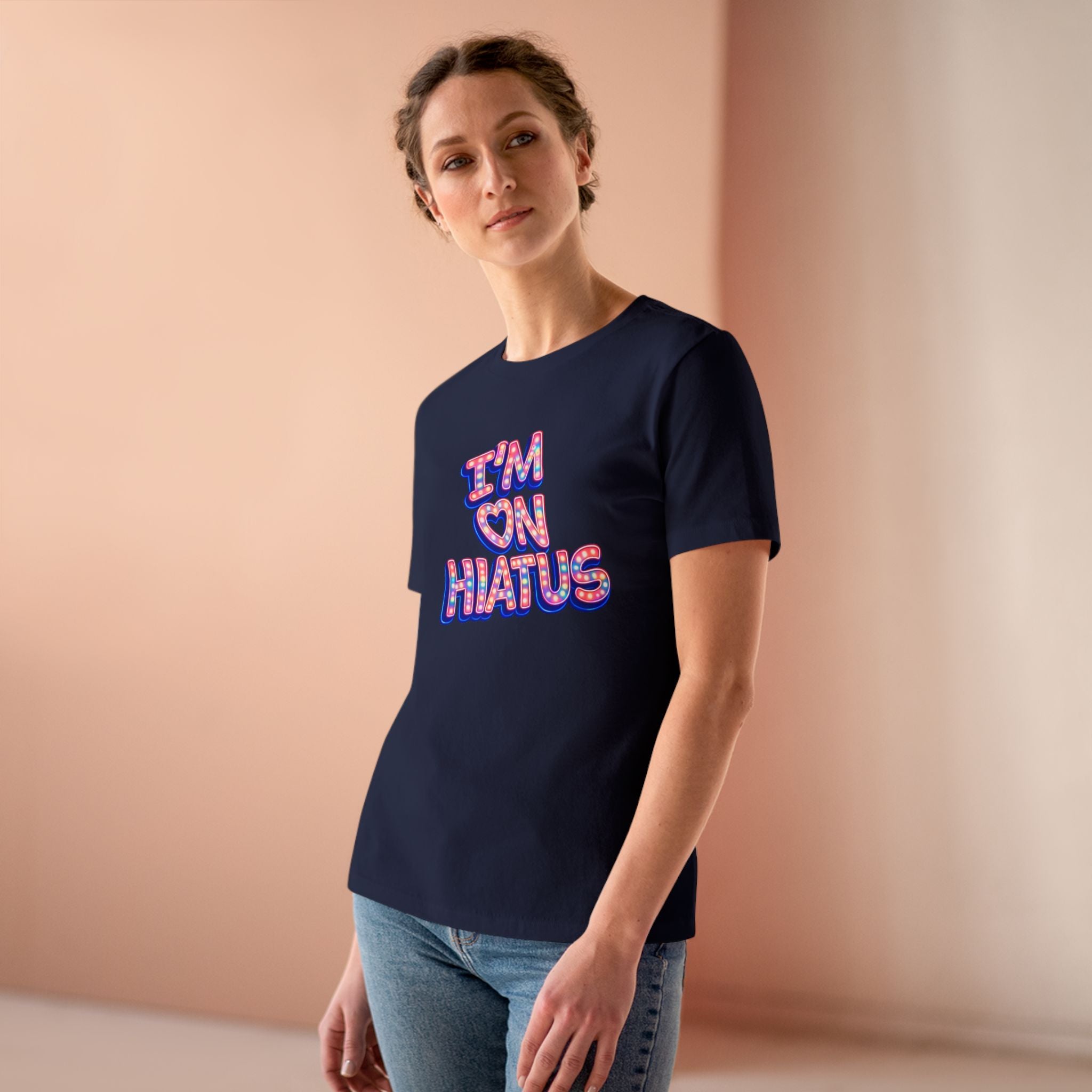 A woman wearing a navy blue t-shirt with a colorful graphic that reads 'I'M ON HIATUS' in a retro font, paired with blue jeans, standing against a soft peach-colored background.
