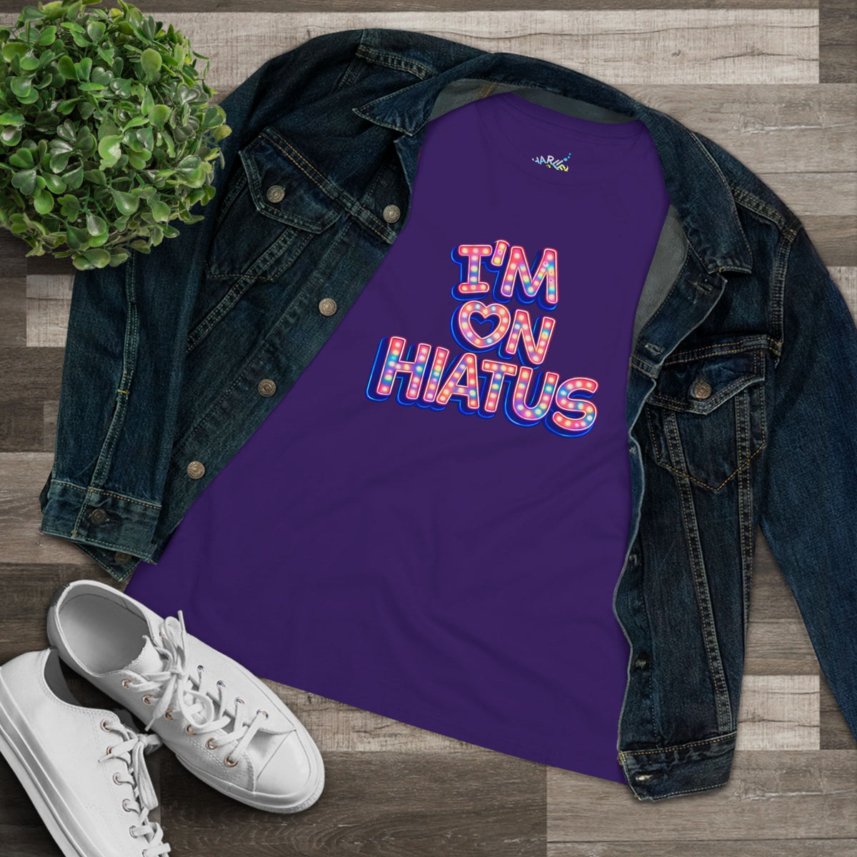 A purple graphic tee with a colorful retro graphic reading 'I'M ON HIATUS' styled in a flat lay with a dark denim jacket, white sneakers, and a small green plant on a wood plank background.