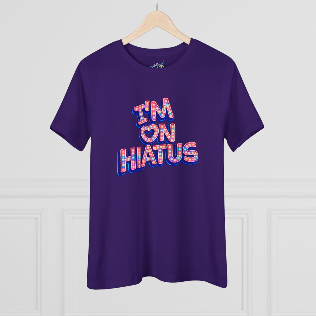 A purple short-sleeve t-shirt hanging on a wooden hanger against a white paneled wall. The shirt features a graphic text design reading 'I'M ON HIATUS' in a bubbly, 3D-style font filled with a red, white, pink,  and blue pattern.