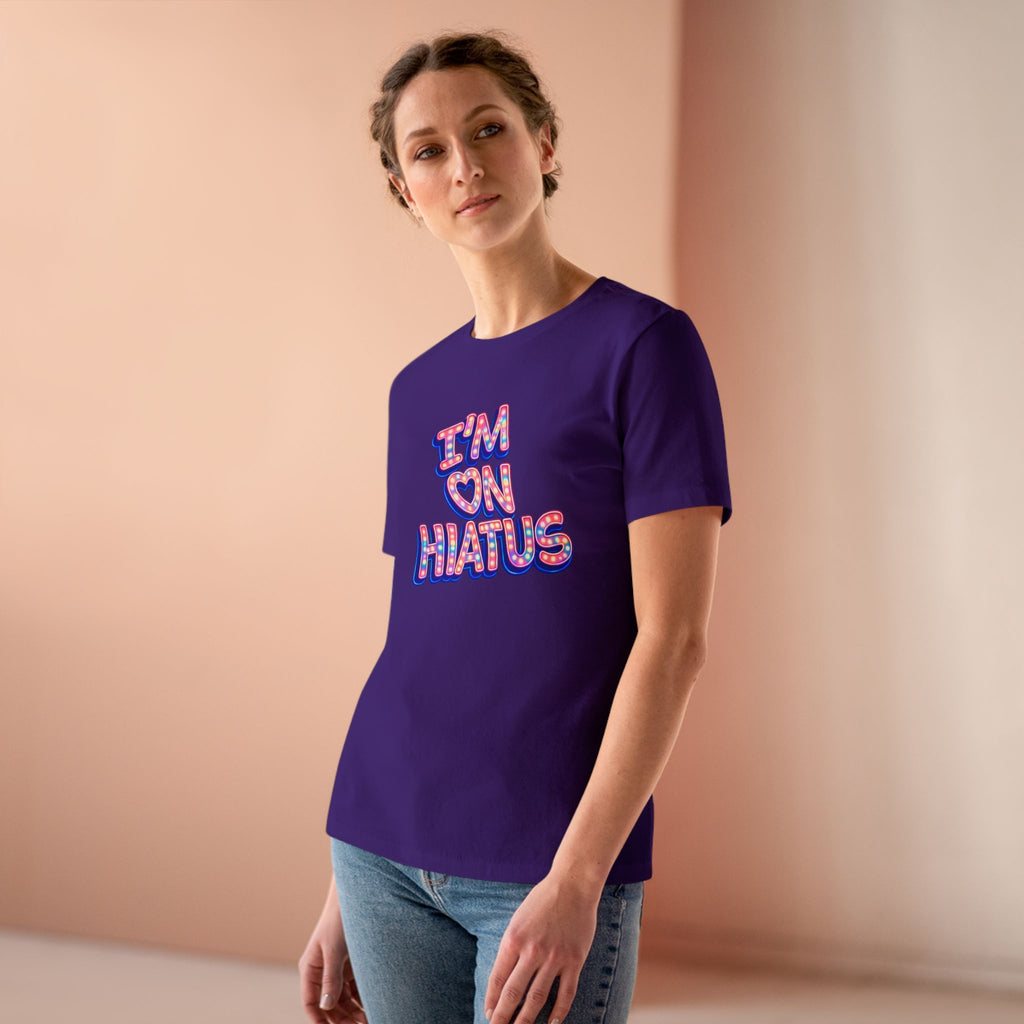 A woman wearing a purple short-sleeve t-shirt with a colorful graphic that reads 'I'M ON HIATUS' in a retro font, paired with blue jeans, standing against a soft peach-colored background.