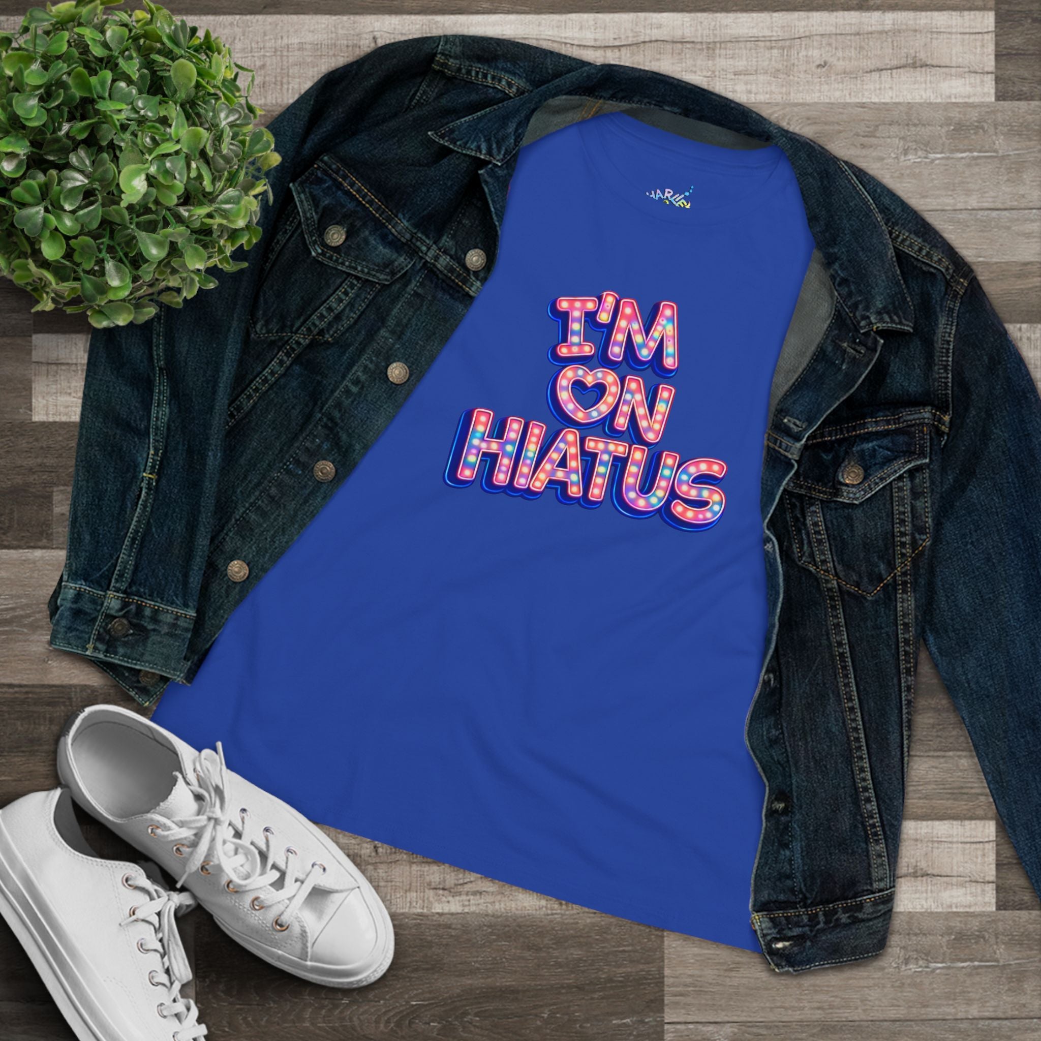 A royal blue tank top with a colorful retro-style graphic reading 'I'M ON HIATUS' with a heart, styled in a flat lay with a dark denim jacket, white sneakers, and a small green plant on a wood plank background.