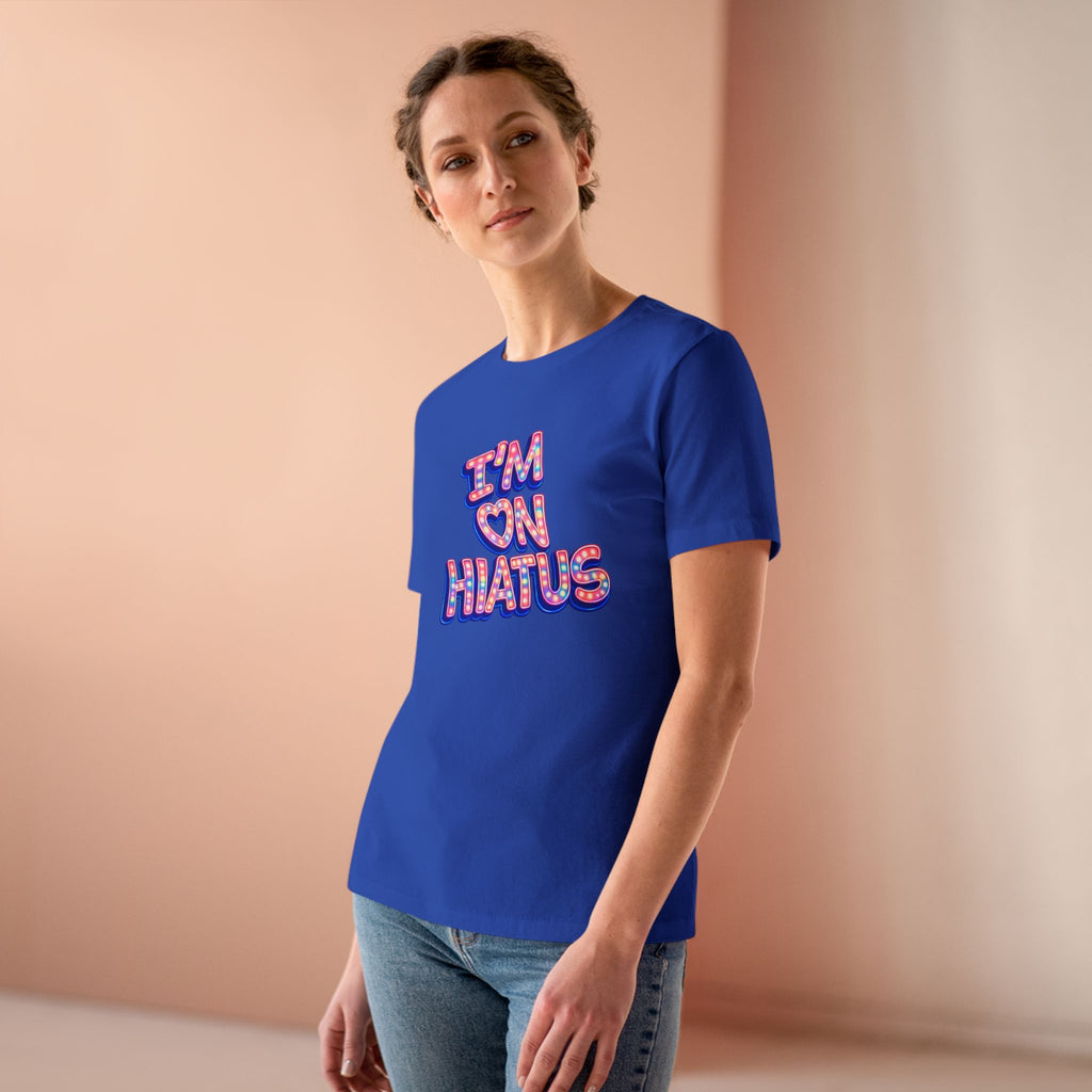 A woman wearing a royal blue t-shirt with 'I'M ON HIATUS' printed on the front in a pink and red patterned font, paired with blue jeans, standing against a soft peach-colored background.