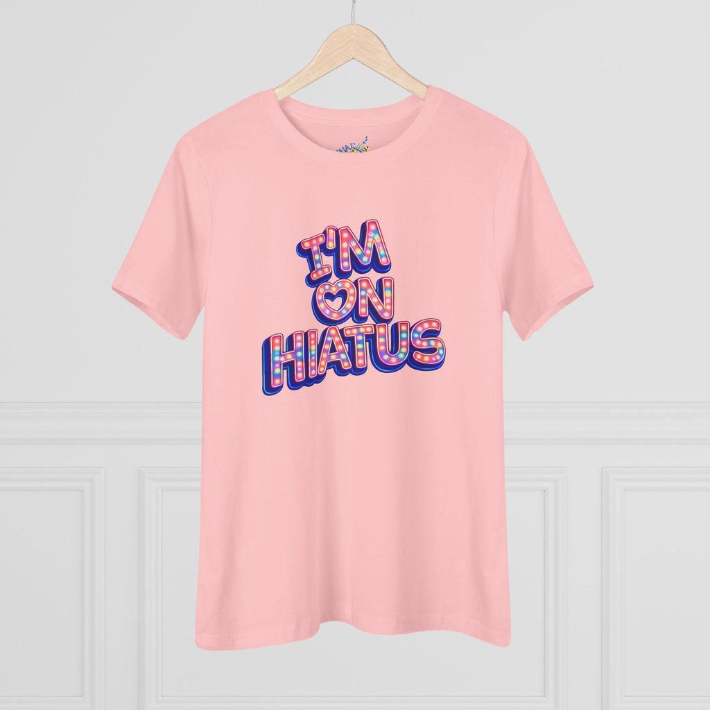 A light pink short-sleeve t-shirt hanging on a wooden hanger against a white paneled wall. The shirt features a graphic text print reading 'I'M ON HIATUS' in a 3D bubble font filled with a red, white, pink, and blue pattern.
