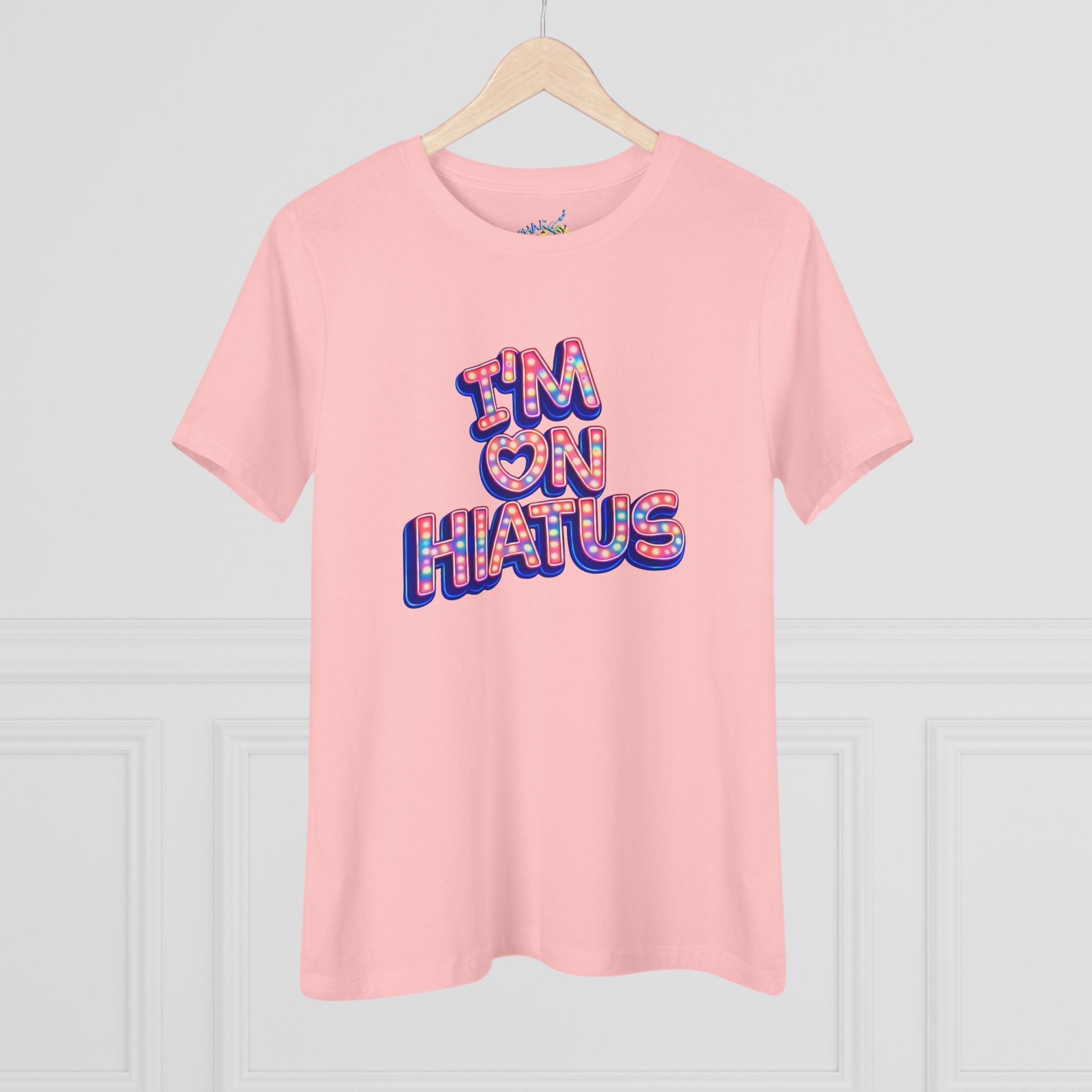 A light pink short-sleeve t-shirt hanging on a wooden hanger against a white paneled wall. The shirt features a graphic text print reading 'I'M ON HIATUS' in a 3D bubble font filled with a red, white, pink, and blue pattern.